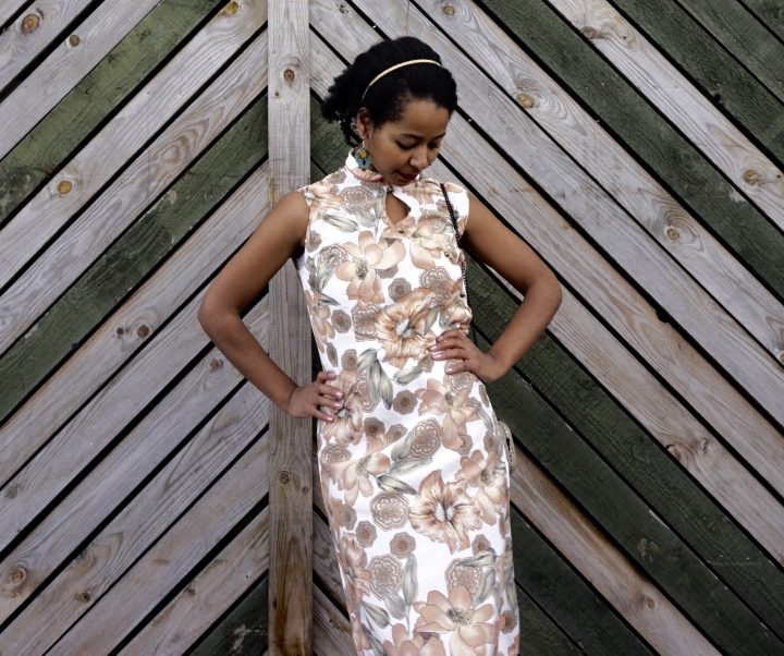 Megan is standing against a wooden fence. She is wearing her handmade qipao. The fabric has a floral print. Megan is looking down at the ground.