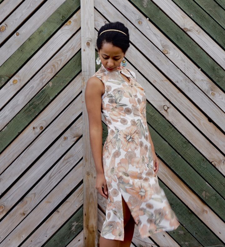 Megan is standing against a wooden fence. She is wearing her handmade qipao. The fabric has a floral print. Megan is looking down at the ground.